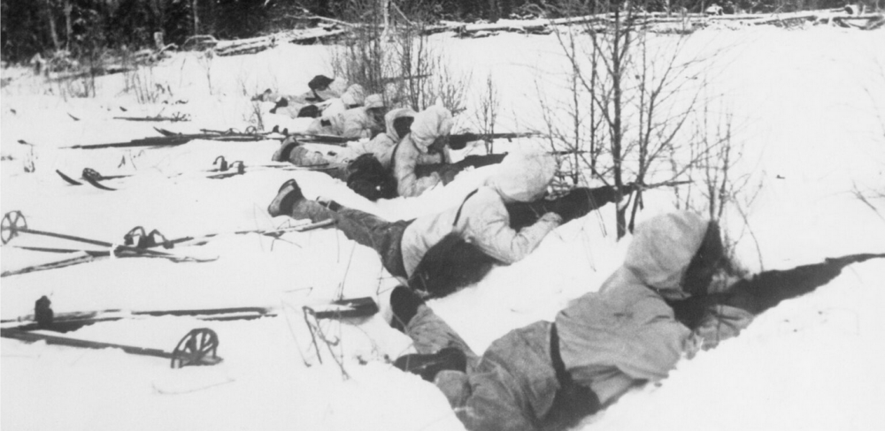 Czarno-biała fotografia przedstawiająca szereg zamaskowanych strzelców przyczajonych za śnieżnym wałem