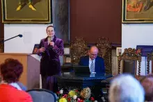 Mężczyzna w ciemnym garniturze siedzi za stołem a kobieta w fioletowej garsonce za mównicą