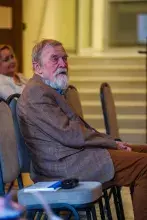 Starszy pan z brodą w brązowej marynarce siedzi na krześle i słucha