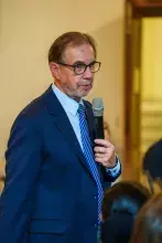 Męzczyzna w ciemnym garniturze i okularach zabiera głos stojąc wśród siedzących uczestników