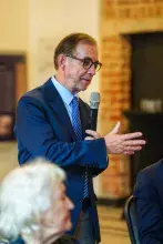 Męzczyzna w ciemnym garniturze i okularach zabiera głos stojąc wśród siedzących uczestników