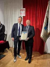 Prof. Cezary Obracht-Prondzyński i Prof. Michał Harciarek Dwóch mężczyzn w garniturach pozuje do zdjęcia, jeden z nich trzyma pamiątkową plakietkę. Stoją na tle biało-czerwonych zasłon i flag.