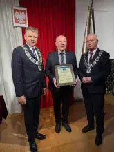 wręczenie rzech mężczyzn w garniturach pozuje do zdjęcia podczas oficjalnej uroczystości. Mężczyzna pośrodku trzyma oprawioną plakietkę, a dwaj mężczyźni po bokach noszą ceremonialne łańcuchy urzędowe. W tle widać czerwone zasłony i polską flagę.