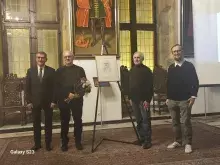 4 mężczyzn stojących na sali z białymi ścianami a między nimi kartka na stelażu dotycząca wystawy.