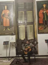 mężczyzna grający na gitarze i siedzący przed wielkim oknem a za nim portrety wiszące na ścianie