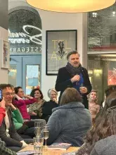 Ujęcie ze spotkania promocyjnego książki. W centrum prof. Mariusz Czepczyński z mikrofonem i kieliszkiem w dłoni, zwracający się do publiczności. Na pierwszym planie, na drewnianym stoliku, leżą egzemplarze książki „Nowa geografia krytyczna”. Wokół siedzą uśmiechnięci uczestnicy wydarzenia, a w tle widoczne są przeszklone drzwi wejściowe do lokalu.