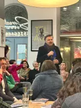 Fotografia przedstawiająca atmosferę spotkania w księgarni-kawiarni. Prof. Mariusz Czepczyński kontynuuje swoją wypowiedź, a zgromadzeni goście, siedzący na kanapach i krzesłach, z uwagą go słuchają. Na stolikach widoczne są napoje oraz egzemplarze nowej publikacji. Wnętrze jest przytulne, z wyraźnym oświetleniem punktowym i dużymi witrynami, za którymi widać wieczorną scenerię.