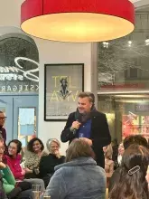 Mężczyzna w średnim wieku (prof. Mariusz Czepczyński) stoi i przemawia do mikrofonu podczas spotkania autorskiego w księgarni. Ma na sobie ciemną marynarkę, niebieską koszulę i gruby, fioletowy szal owinięty wokół szyi. W tle widać siedzących słuchaczy, plakaty na ścianach oraz duże okno z neonowym napisem „Księgarnia”. Nad prelegentem wisi duża, okrągła lampa dająca ciepłe światło.