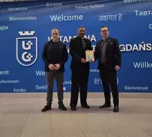 Trzej mężczyźni w strojach biznesowych stoją wewnątrz nowoczesnego budynku na tle dużej, jasnoniebieskiej ściany z napisem „WITAMY NA UNIWERSYTECIE GDAŃSKIM” oraz słowem „Welcome” w wielu językach. Mężczyzna w środku trzyma egzemplarz książki z żółtą okładką. Po lewej stronie widoczne jest logo Uniwersytetu Gdańskiego.