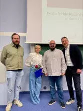 Zdjęcie przedstawiające 4 osoboy podczas wręczenia nagrody, 3 mężczyzn w strojach formalnych i kobieta w stroju formalnym trzymająca dyplom i czerwoną różę