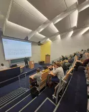 Szerokie ujęcie sali wykładowej z perspektywy górnych rzędów. Na dole, przy mównicy, stoi prelegent w ciemnym garniturze, omawiając slajd dotyczący działań wojskowych i bezpieczeństwa w regionie Cabo Delgado. W ławkach siedzi grupa słuchaczy skupiona na prezentacji.