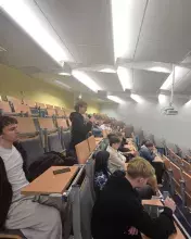 Perspektywa z boku rzędów w sali wykładowej. Młoda osoba w okularach i czarnej bluzie stoi w ławce, trzymając mikrofon i zadając pytanie podczas sesji Q&A. Inni studenci siedzą obok, słuchając wypowiedzi.