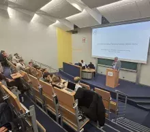 Sala wykładowa. Starszy mężczyzna w jasnej marynarce stoi przy mównicy z mikrofonem, prawdopodobnie wprowadzając prelegenta lub prowadząc dyskusję. W tle na ekranie widoczny tytuł wykładu o polityce Rwandy. W ławkach siedzą uczestnicy spotkania.