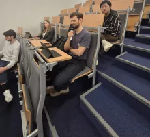 Zbliżenie na widownię w nowoczesnej, amfiteatralnej sali wykładowej. Mężczyzna w fioletowym t-shircie siedzi w ławce i zadaje pytanie do mikrofonu. Obok niego inni studenci korzystają z tabletów i laptopów.