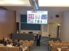 Zdjęcie przedstawiające prelegenta w trakcie wykładu mówiącego do grupy studentów i uczniów