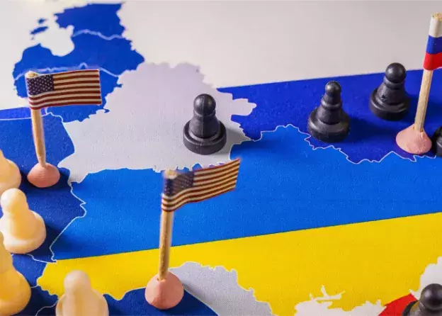 zdjęcie mapy Ukrainy w barwach ukraińskeij flagi; wokół granic ustawione pionki: czarne (Rosja, Białoruś) i białe (państwa Zachodu); małe flagi Rosji, WUSA i UE