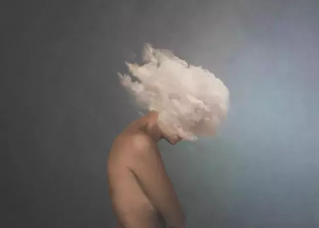 Obraz kobiety (od klatki piersiowej w górę, widok z boku) pochylającej głowę otoczoną chmurami, na ciemnoszarym tle