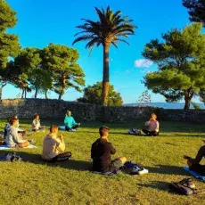 Grupa medytujących młodych ludzi na trawiastym skwerze w słońcu, w tle pojedyncze drzewa i palmy